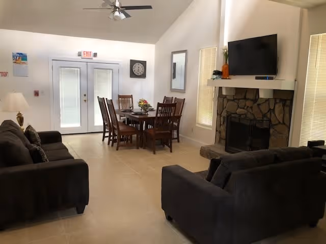 A bright living room and dining area with two dark sofas facing each other, a stone fireplace with a mounted flat-screen TV above it, a dining table with six wooden chairs, a ceiling fan, and double glass doors with blinds. The room has light-colored tile flooring and white walls with windows on either side of the fireplace.