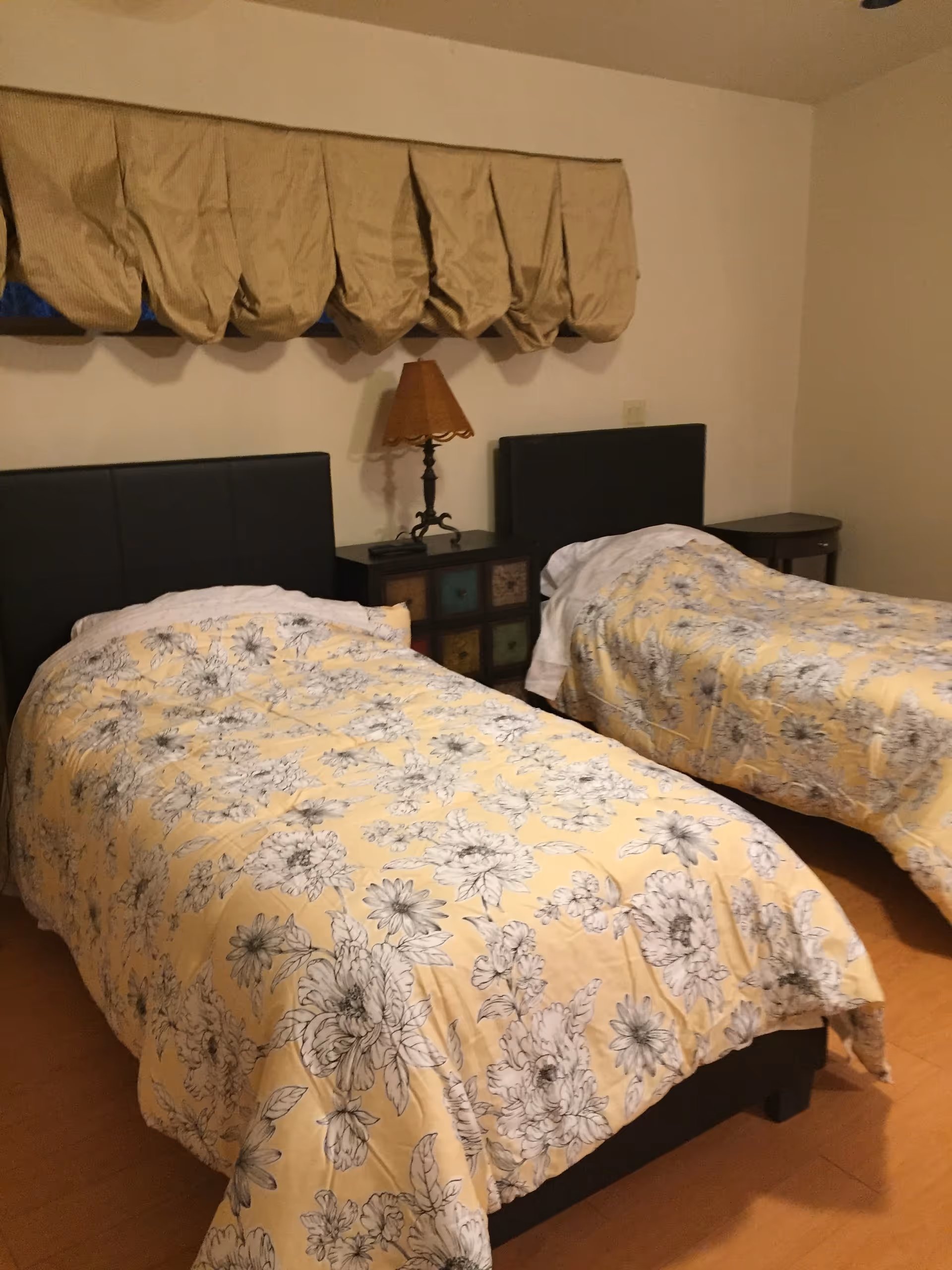 A bedroom with two twin beds dressed in yellow floral comforters, a nightstand with a lamp between them and a window valance above.