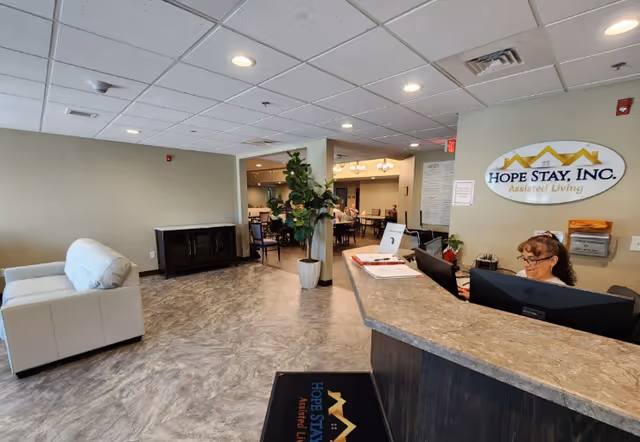 Reception area of Hope Stay Memory Care facility with a woman working behind the front desk, a white couch on the left, a potted plant near the center, and a dining area visible in the background.