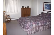Furnished bedroom with a bed, wooden dresser, rocking chair, and vertical blinds on the window.