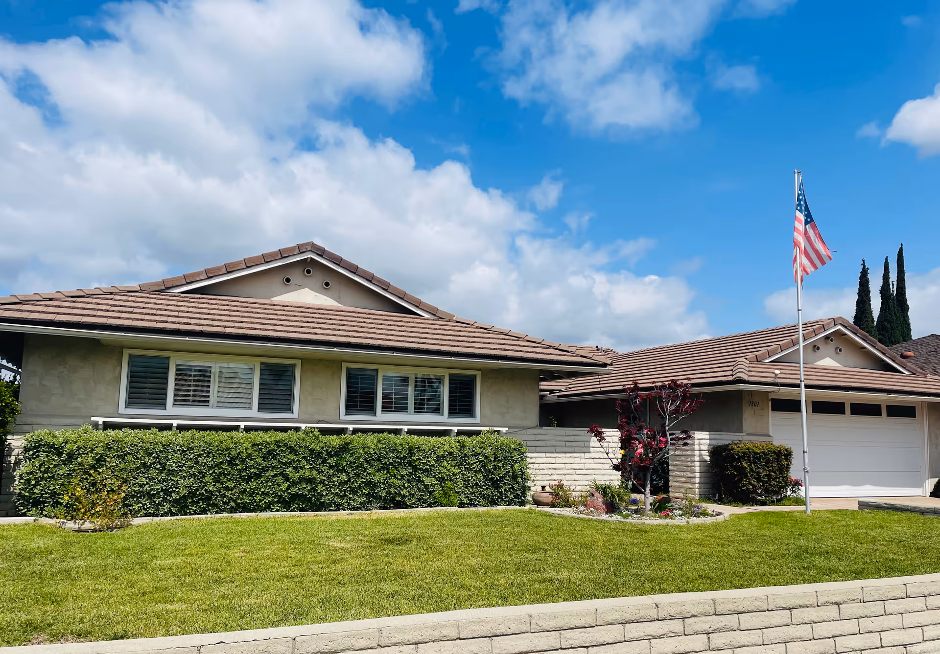 Single-story residential building with a tiled roof, beige exterior walls, and multiple windows with shutters. The front yard has a well-maintained green lawn, bushes, and a small tree. An American flag is mounted on a flagpole near the driveway under a partly cloudy blue sky.