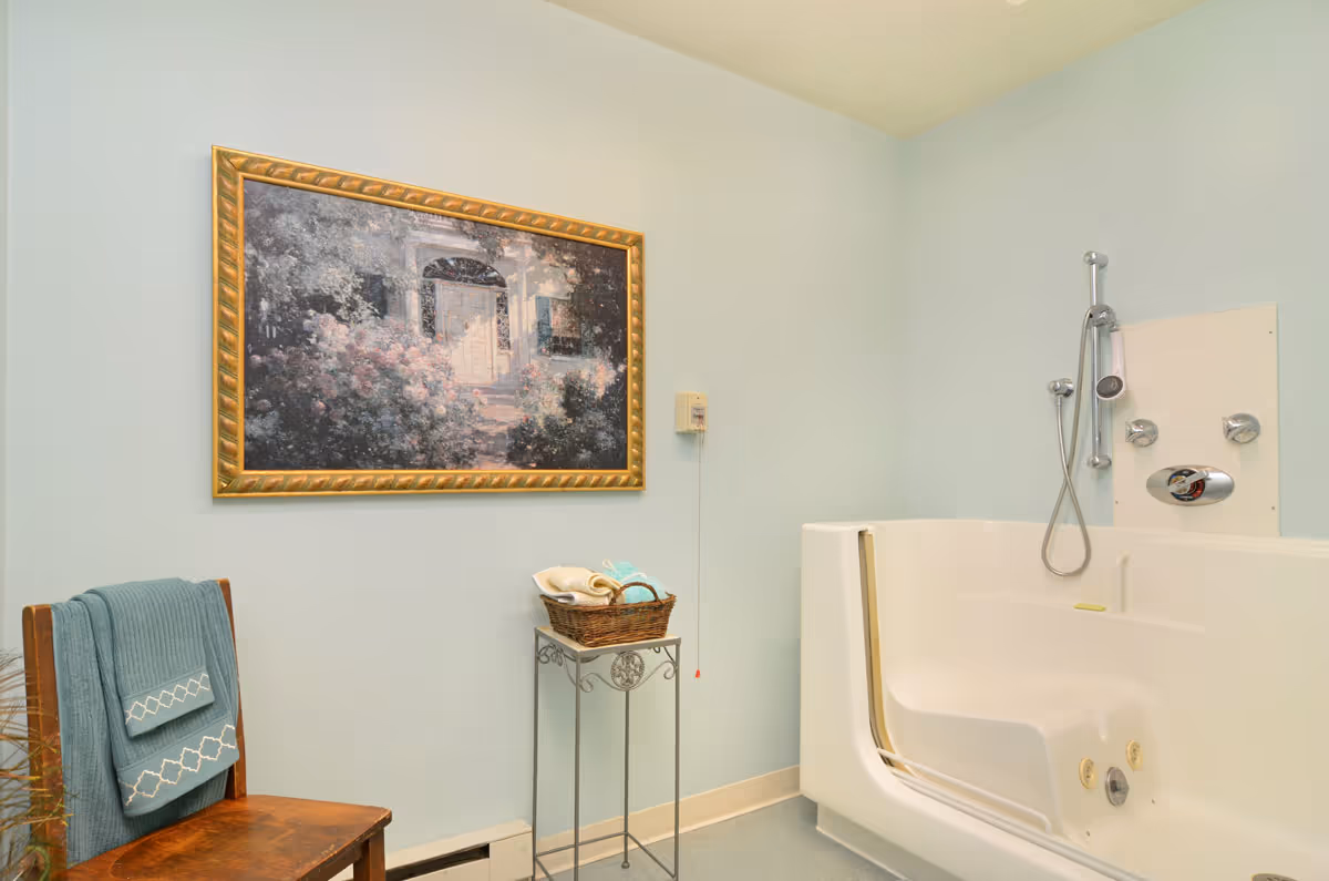 Light-blue bathroom featuring a white walk-in bathtub with handheld shower, a framed painting on the wall, a chair with towels, and a small table with a basket.