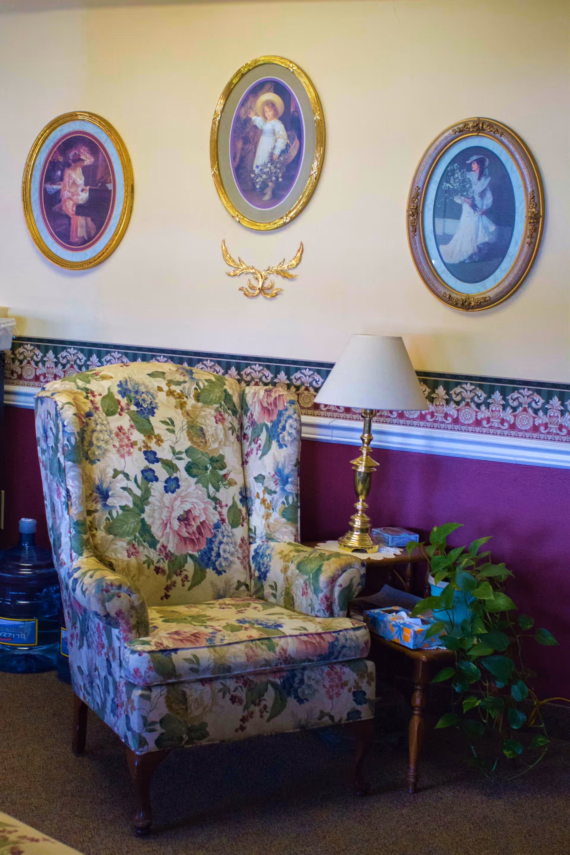 Floral upholstered wingback chair beside a small side table with a lamp and a potted plant beneath three oval framed portraits on a decorated wall.