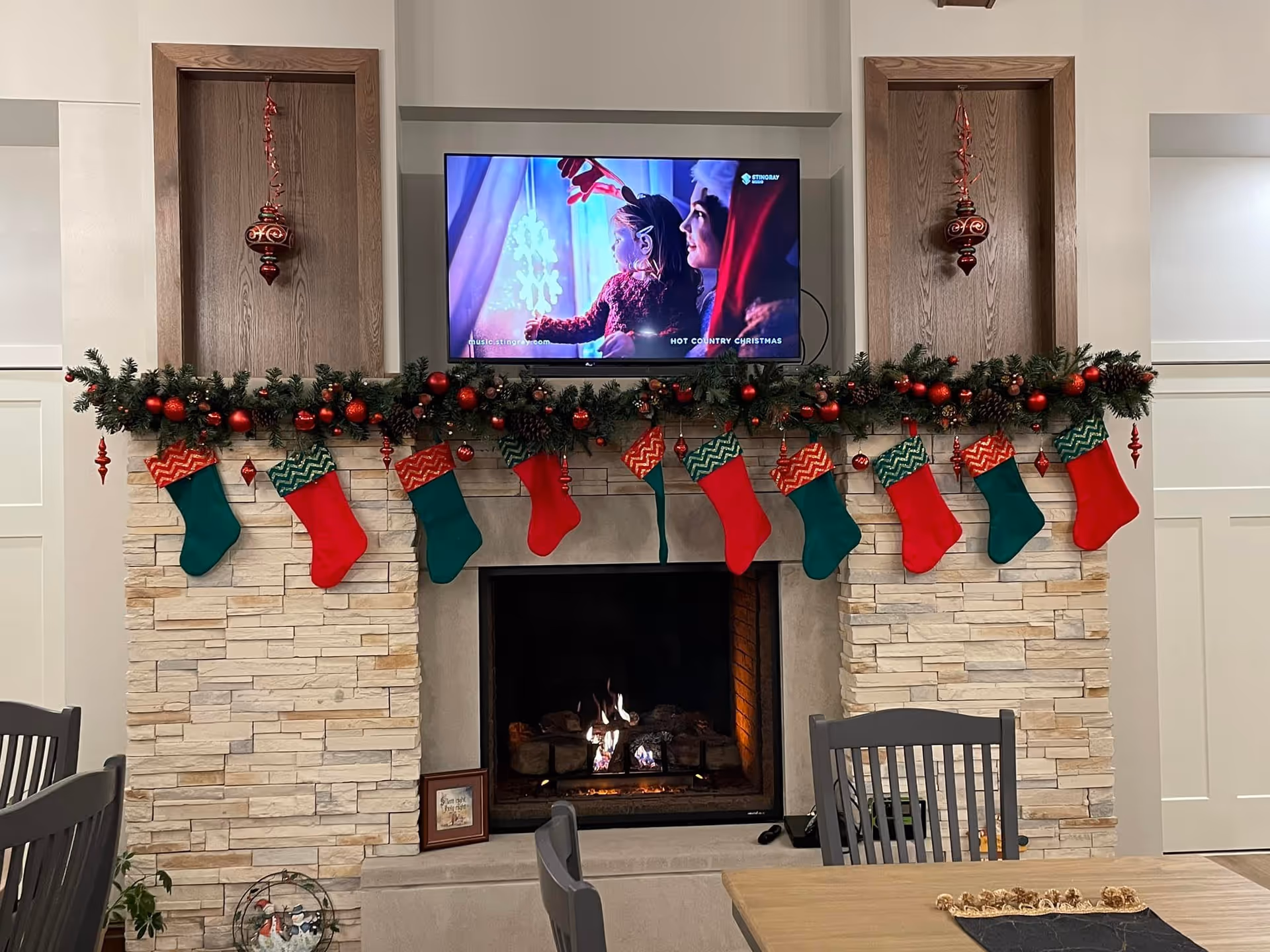 A cozy living room area decorated for Christmas with a stone fireplace. Above the fireplace is a mounted flat-screen TV showing a holiday scene with a child and an adult wearing a Santa hat. The mantel is adorned with green garland, red ornaments, and nine red and green Christmas stockings hanging in a row. Two wooden panels flank the TV, each with a red ornament hanging. In front of the fireplace are gray chairs and a wooden table with a decorative centerpiece.