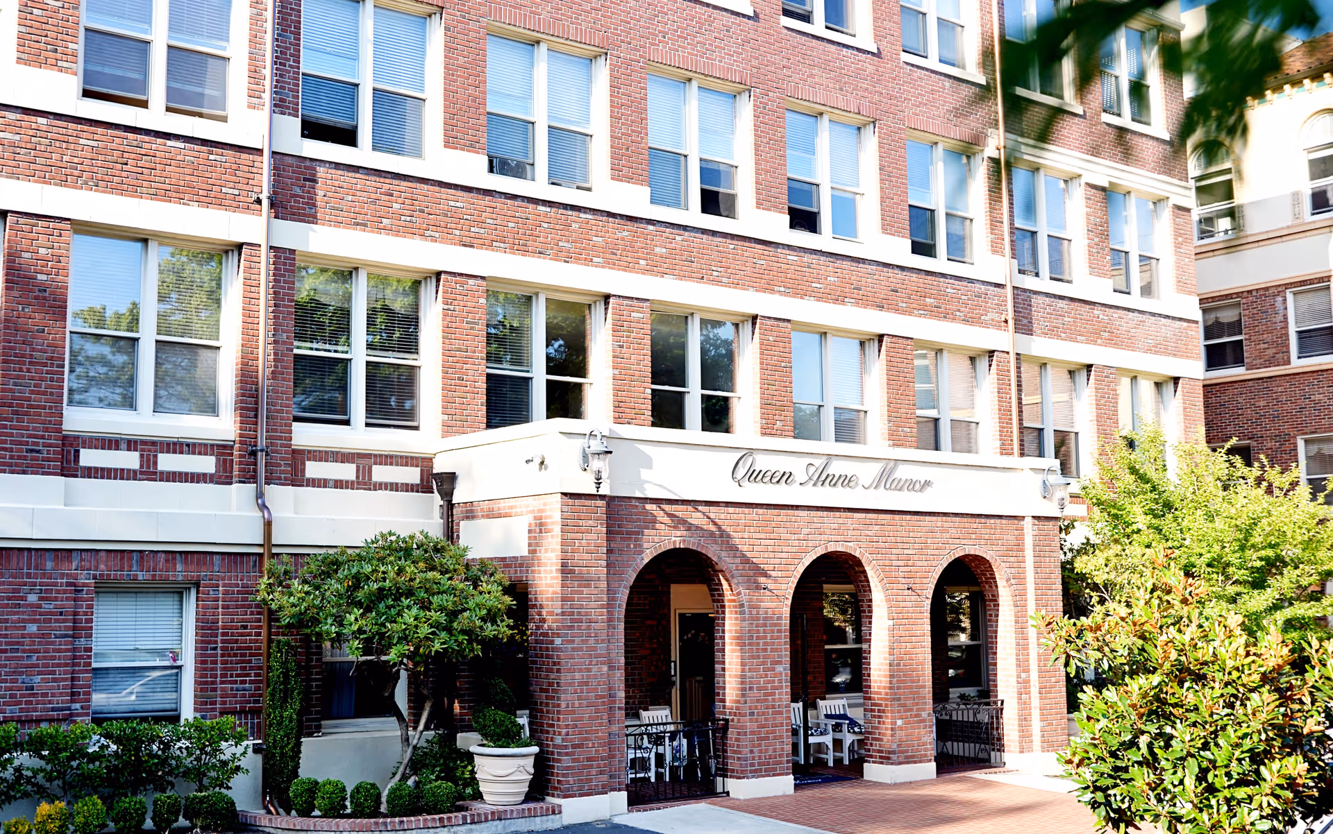 Brick multi-story building front with an arched entrance labeled "Queen Anne Manor" and small landscaped planters.