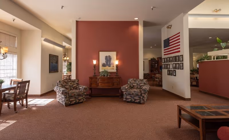 A spacious senior living common area with two patterned armchairs facing a wooden sideboard against a red accent wall. The sideboard has two lit lamps and a framed picture above it. To the left, there is a dining area with wooden chairs and tables near large windows with blinds. On the right wall, an American flag and framed photographs are displayed. The room has carpeted floors and soft lighting.