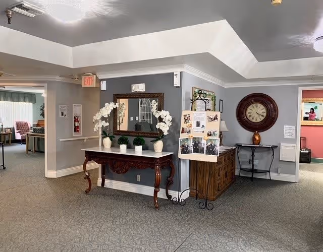 Interior view of a senior living facility hallway with a decorative wooden table holding white orchid plants and small green topiary balls. Above the table is a large framed mirror. To the right, there is a wooden cabinet with a large round wall clock above it and a black metal chair. A display board with photos is placed on a stand next to the cabinet. The hallway leads to other rooms, one of which has a pink wall and a person visible in the background.