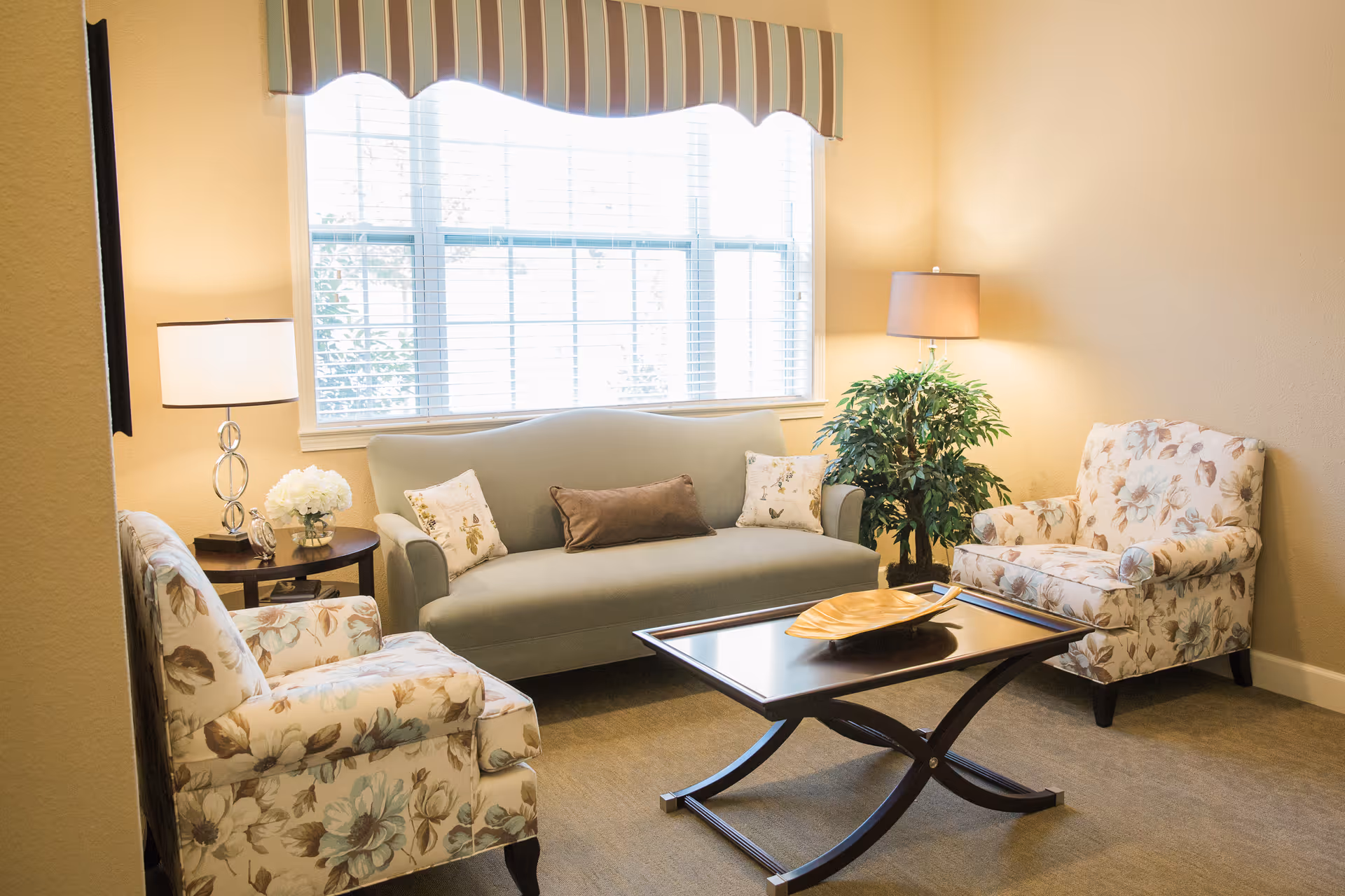 A cozy living room with a large window covered by a striped valance. The room features a light green sofa with three decorative pillows, two floral-patterned armchairs, a dark wooden coffee table with a decorative tray, a side table with a lamp and flowers, a floor lamp, and a potted plant in the corner.