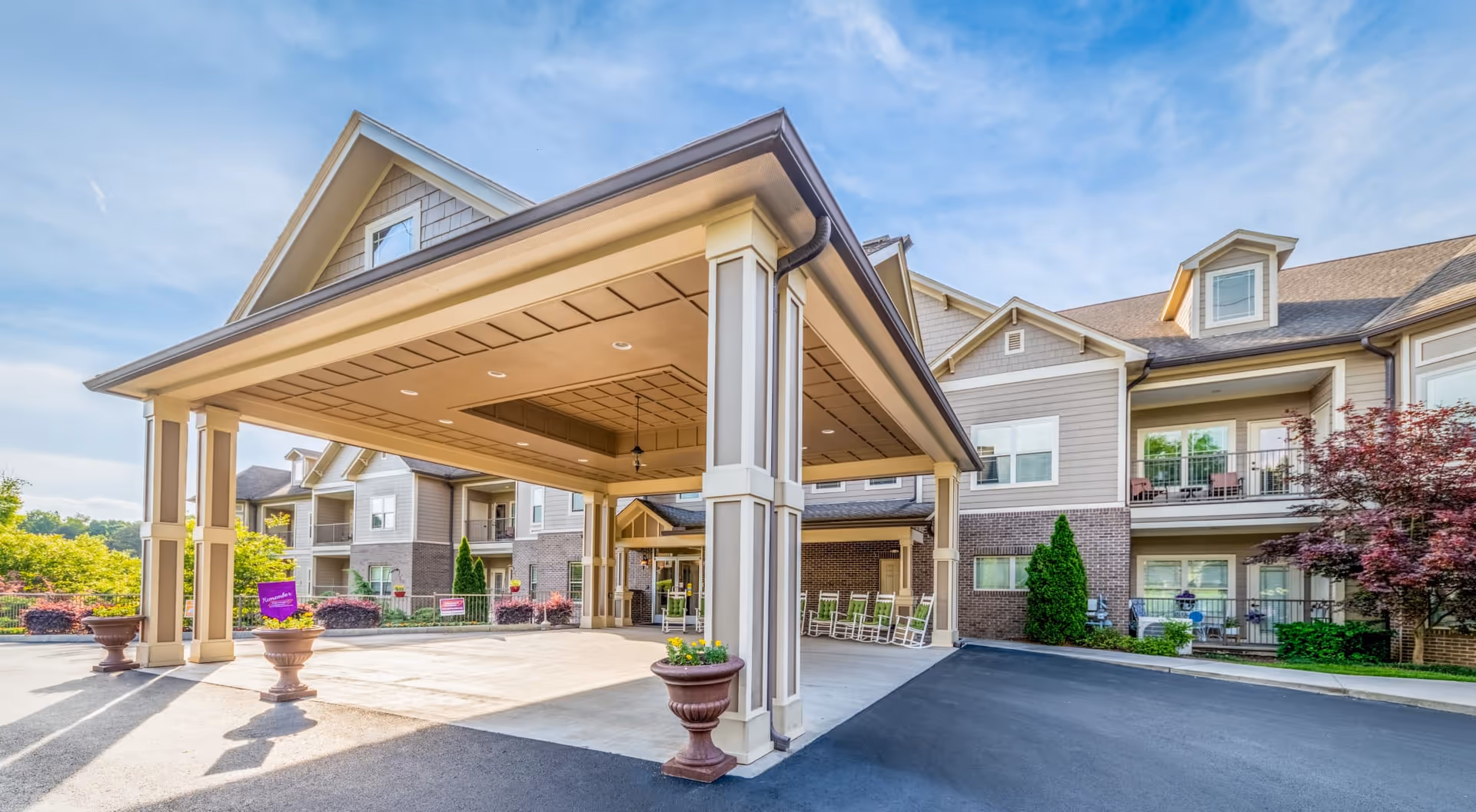 Exterior view of a senior living facility named Rain Forest Landings featuring a covered entrance with columns, rocking chairs on the porch, landscaped greenery, and a clear blue sky.