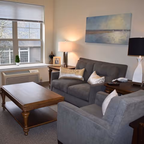 A cozy living room with a gray sofa and armchair, wooden coffee table, side tables with lamps, wall art, and a window.