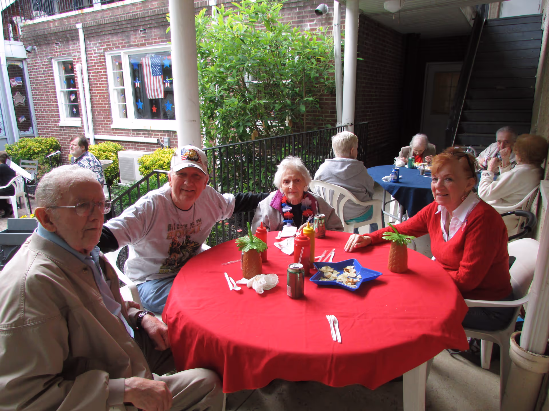 A group of elderly people sitting around a round table covered with a red tablecloth on a covered outdoor patio. The table has two pineapple-shaped decorations, some soda cans, condiments, and a blue star-shaped plate with snacks. In the background, more elderly people are seated at another table with a blue tablecloth. The setting is surrounded by brick walls and greenery.