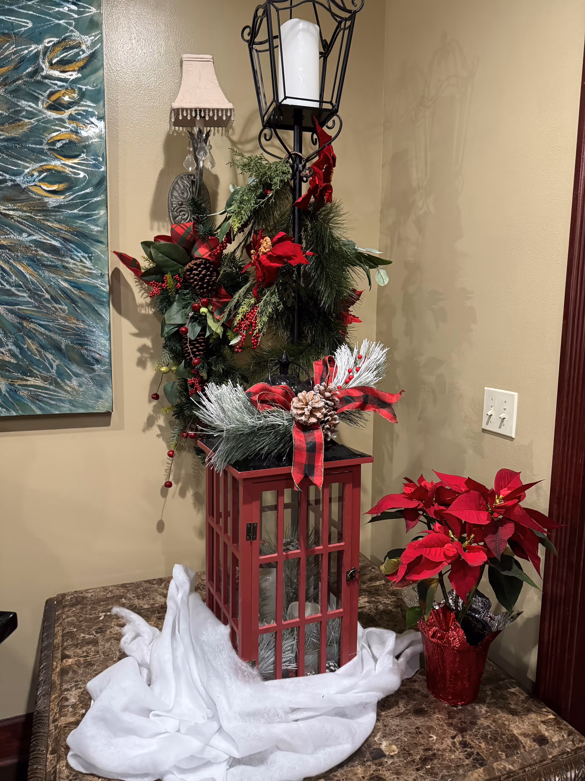 A festive holiday decoration setup on a marble table featuring a red lantern adorned with pine branches, pinecones, and a red and black plaid ribbon. Next to the lantern is a red poinsettia plant in a red foil-wrapped pot. The background includes a beige wall with a light switch, a decorative wall sconce with a small lampshade, and part of a blue and gold abstract painting.