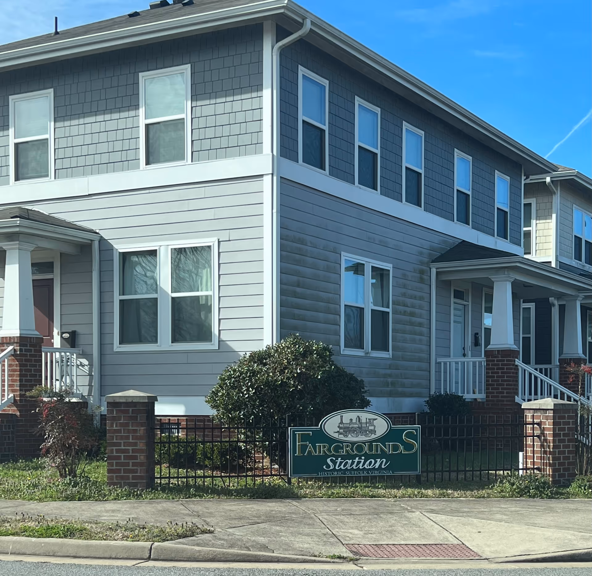 Exterior view of a two-story residential building with gray siding and white trim. There is a small fenced yard with bushes and a sign that reads 'Fairgrounds Station Historic Suffolk Virginia' in front of the building. The sky is clear and blue.