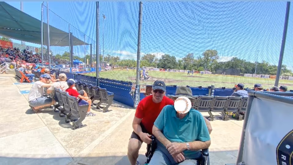 An outdoor baseball stadium with spectators seated on benches and bleachers. In the foreground, a man in a red shirt and cap is crouching next to an elderly person in a wheelchair wearing a teal shirt and hat. The field is visible through a protective netting, with players and trees in the background under a clear blue sky.
