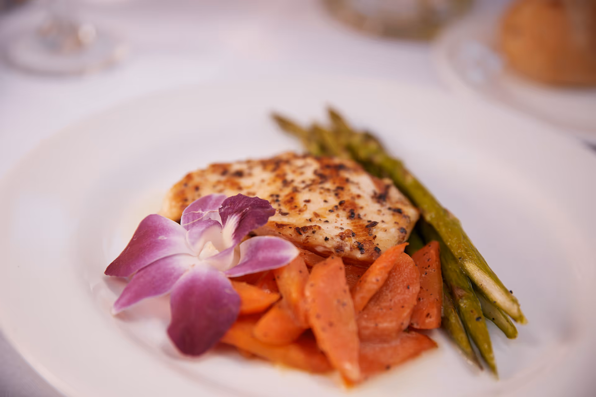 A plated meal featuring a grilled chicken breast, cooked asparagus, sliced carrots, and a purple orchid flower garnish on a white plate.