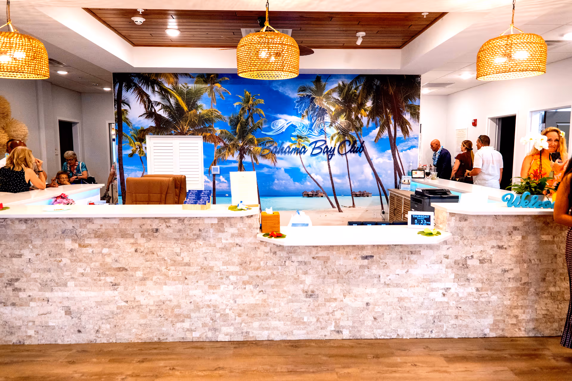 Reception area of Bahama Bay Club Luxury Senior Living with a stone-textured front desk, tropical beach mural with palm trees on the back wall, and several people interacting around the desk. The ceiling has wooden panels and hanging wicker light fixtures.