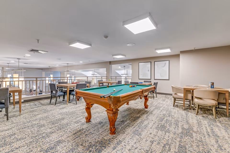 A spacious recreational room with a green felt pool table in the center, surrounded by several tables and chairs. The room has a carpeted floor, beige walls, and modern ceiling lights. Two framed abstract artworks hang on the wall, and the area overlooks a lower level with large windows and hanging pendant lights.