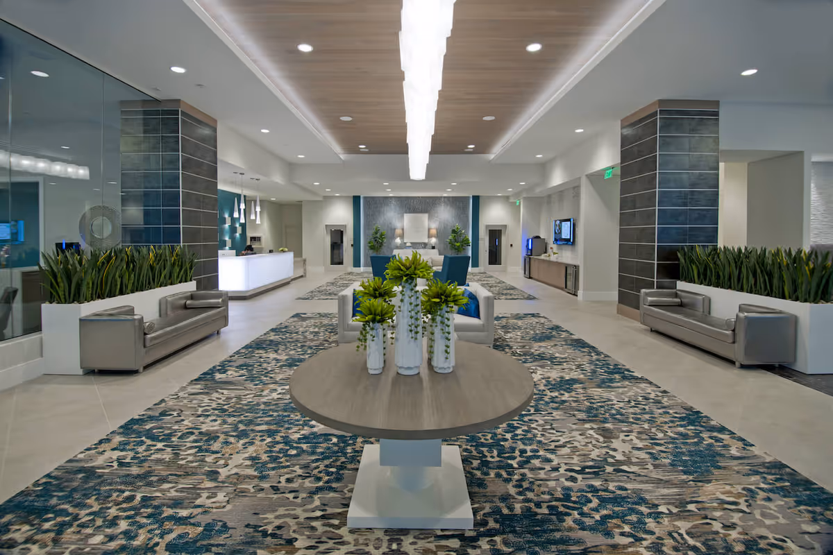 Spacious modern lobby with seating, a central round table of plants, patterned rugs and a reception desk in the background.
