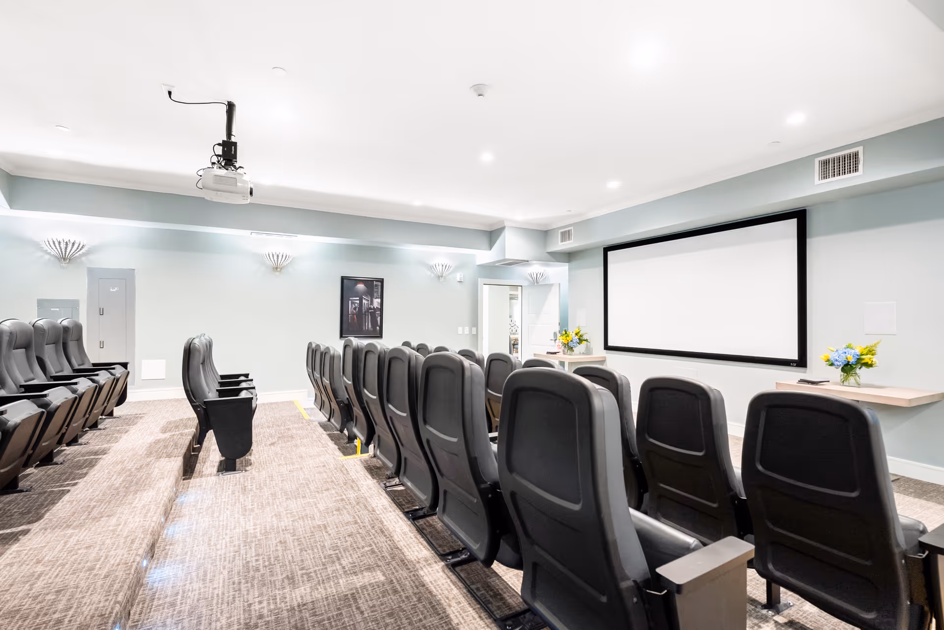 A small theater room with two rows of black cushioned seats facing a large blank projection screen on a light blue wall. There is a ceiling-mounted projector and two small tables with flower arrangements on either side of the screen. The room is well-lit with recessed lighting and wall sconces.