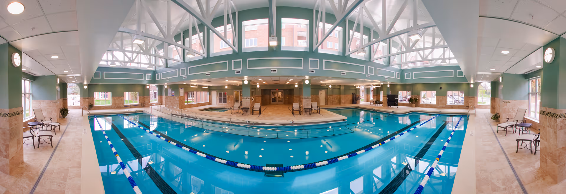 Indoor swimming pool area with clear blue water, lane dividers, and a hot tub in the background. The space features large windows, green walls with decorative trim, beige tiled floors, and several chairs and tables along the sides. The ceiling has white beams and recessed lighting.