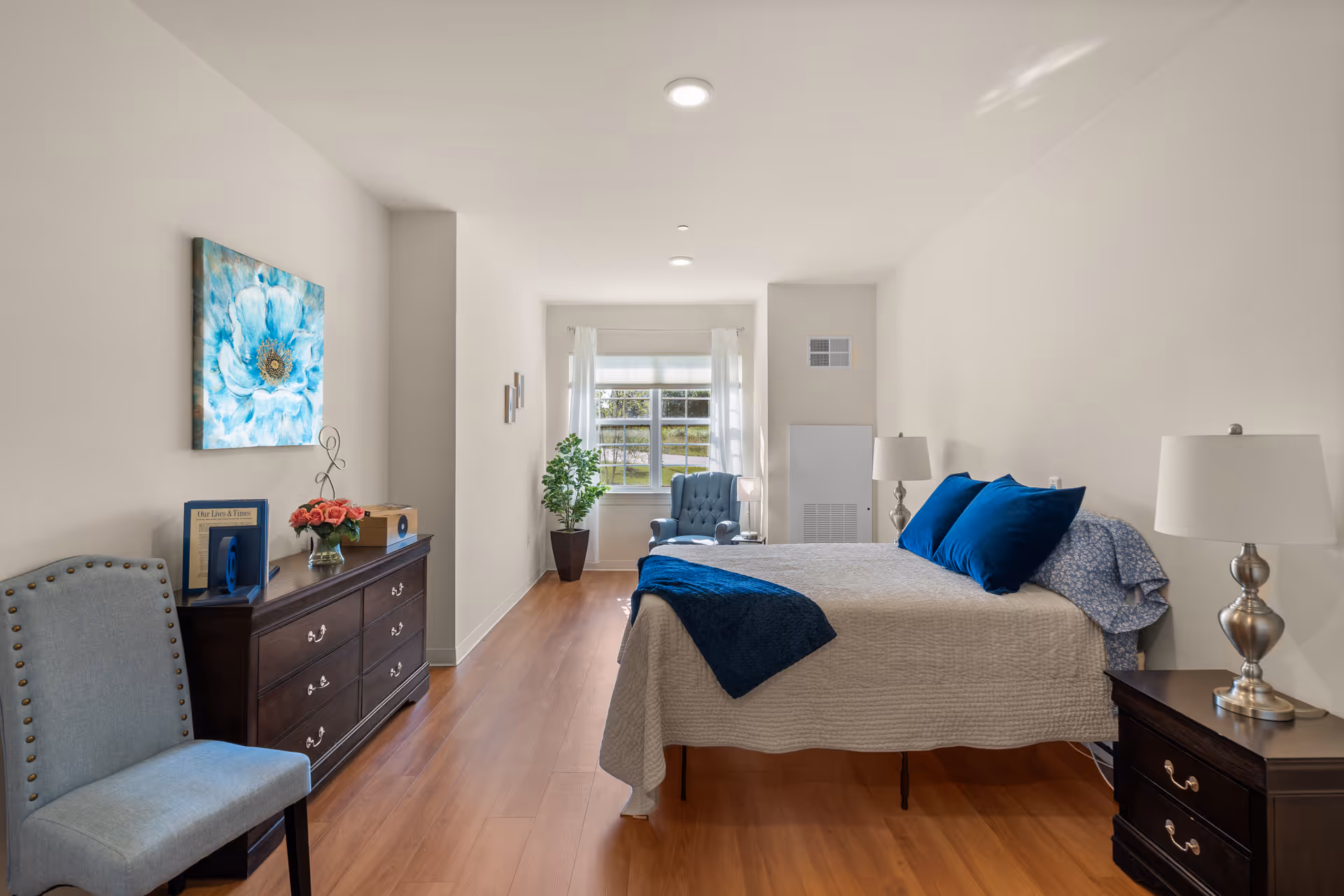 A bright and neatly arranged bedroom in a memory care community featuring a bed with blue pillows and a beige quilt, two bedside tables with lamps, a wooden dresser with decorative items, a blue upholstered chair, a large window with white curtains, and a potted plant near the window.