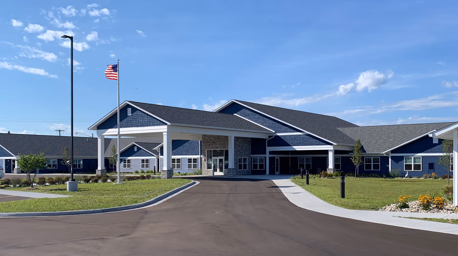 Exterior view of Besser Senior Living Community building with a covered entrance, an American flag on a flagpole, a paved driveway, green lawn, and a clear blue sky with some clouds.