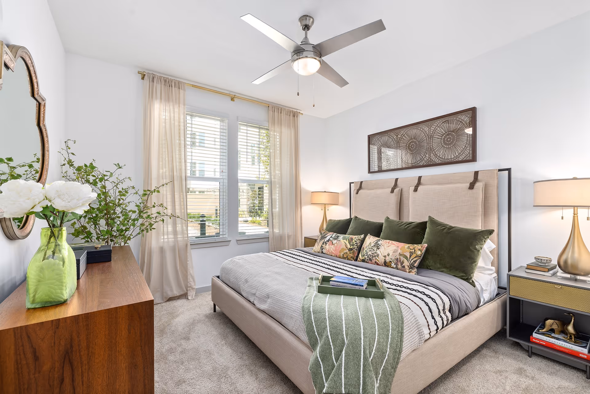 A bright and cozy bedroom with a large beige upholstered bed adorned with green and floral pillows and a striped blanket. There is a wooden dresser with a green vase holding white flowers and greenery, a decorative mirror above it, and two windows with beige curtains letting in natural light. A ceiling fan with a light fixture hangs above, and two bedside tables with lamps flank the bed.