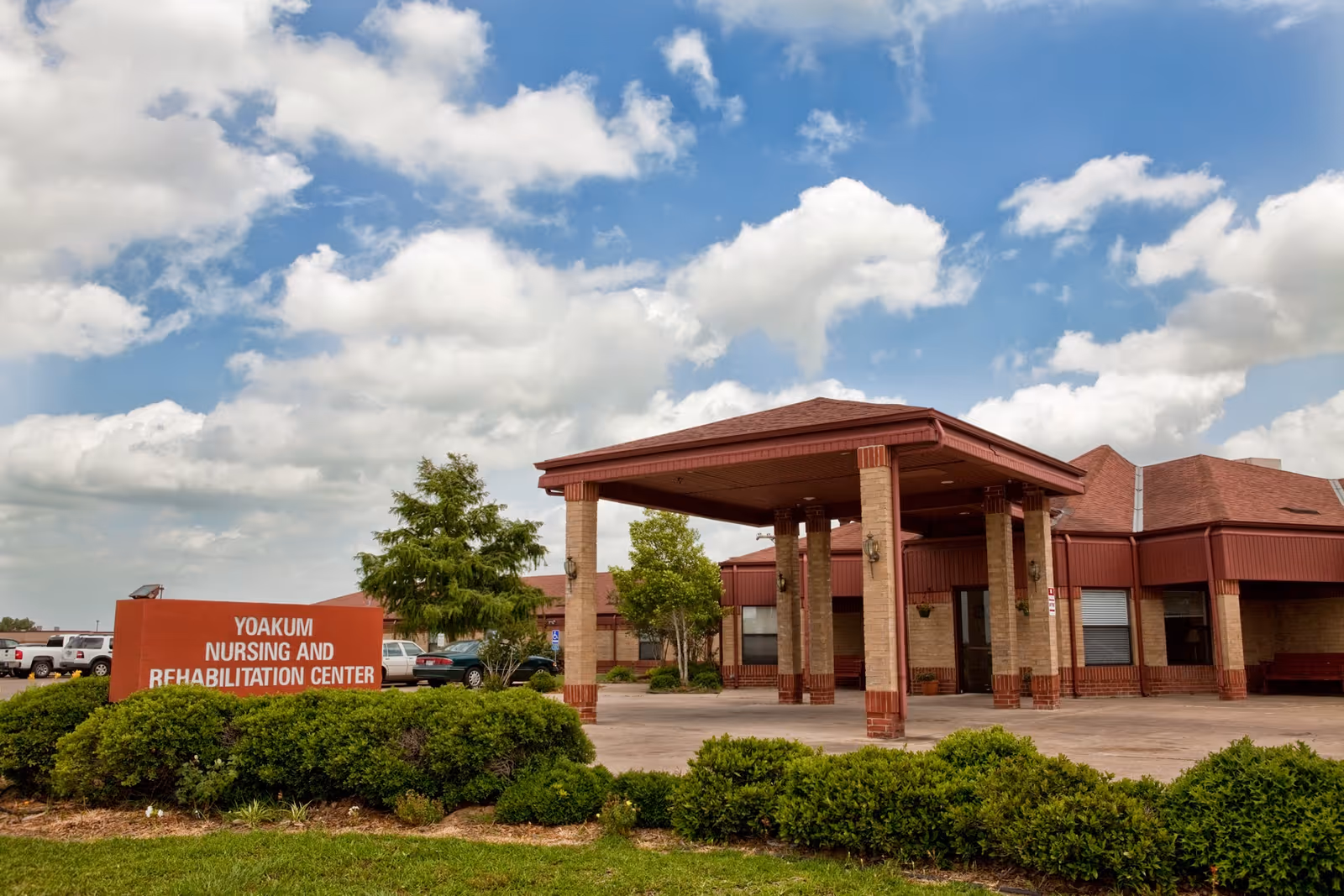 Exterior view of Yoakum Nursing and Rehabilitation Center building with a covered entrance, surrounded by green bushes and trees under a partly cloudy sky.