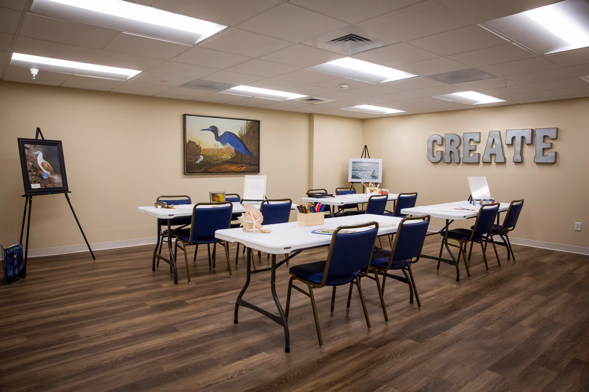 A well-lit activity room with several tables and blue chairs arranged for group activities. The walls are beige, decorated with framed bird paintings and large metallic letters spelling 'CREATE'. Art supplies are placed on the tables, suggesting a creative or craft space.