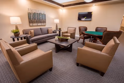 A cozy senior living common area with a gray sofa, four beige armchairs arranged around a wooden coffee table, two side tables with lamps and plants, a wall-mounted TV, and a card table with four chairs in the background. The room has beige walls and carpeted flooring.