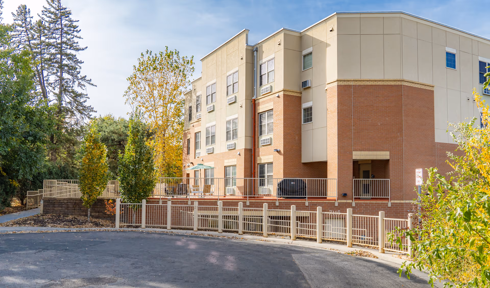 Exterior view of a multi-story senior living facility building with beige and brick facade, surrounded by trees with autumn foliage and a paved driveway in front.