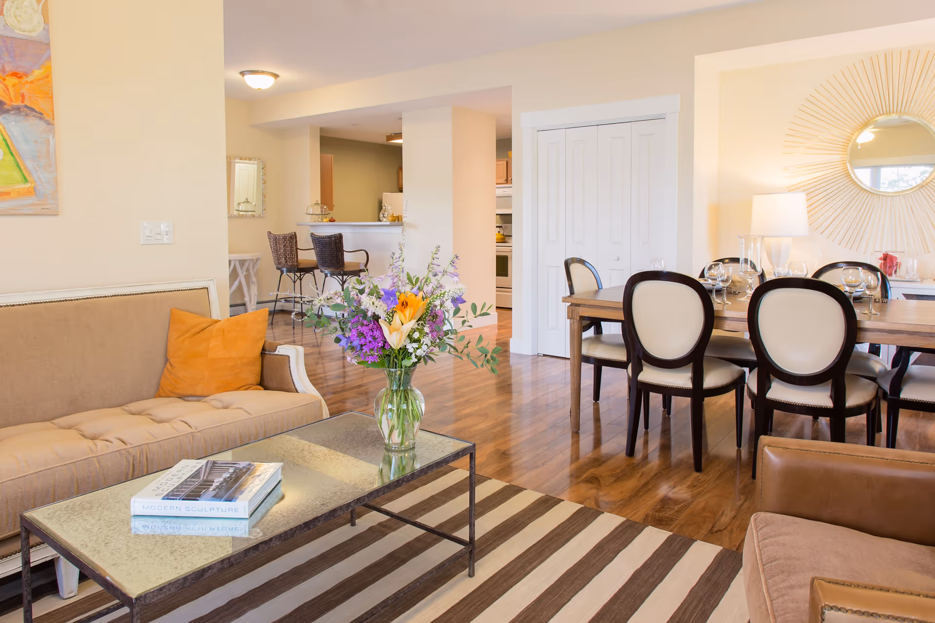 A bright and spacious living and dining area featuring a beige sofa with an orange pillow, a glass coffee table with books and a vase of colorful flowers, a wooden dining table set with six chairs and glassware, a large decorative sunburst mirror on the wall, and a kitchen area with bar stools in the background.