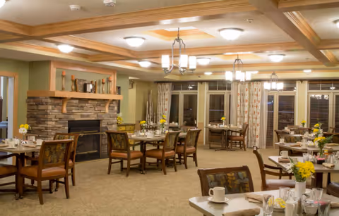 A cozy dining room in a senior living facility with multiple tables set for meals. Each table has chairs, tableware, and small flower arrangements. The room features a stone fireplace with a wooden mantel, warm lighting from ceiling fixtures, and large windows with patterned curtains.