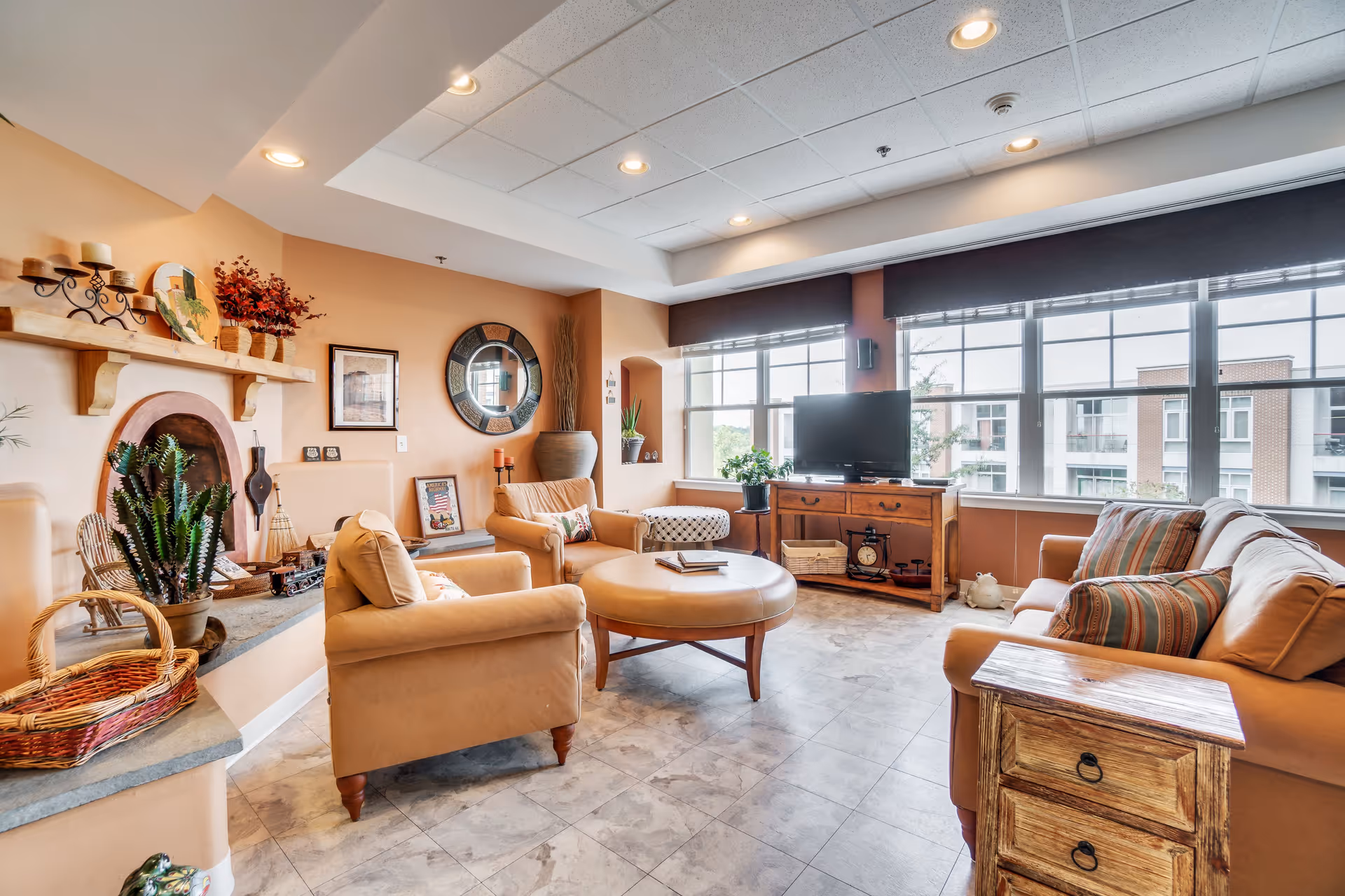 A cozy living room with beige armchairs and a sofa adorned with striped pillows. A round leather ottoman sits in the center on a tiled floor. The room features a wooden TV stand with a flat-screen TV, large windows with black blinds letting in natural light, and decorative elements including plants, a round wall mirror, framed pictures, and candles on a wooden mantel.
