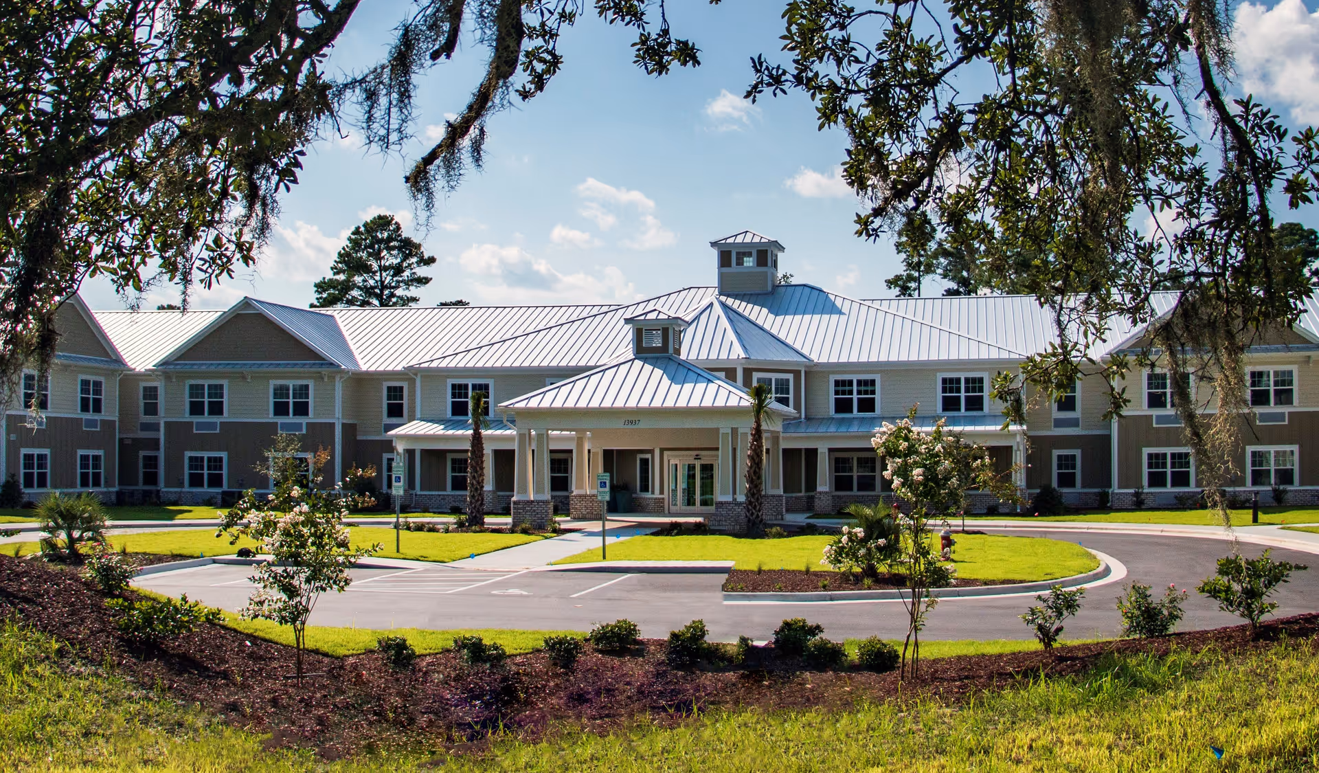 Front exterior view of Arbor Landing at Hampstead, a two-story senior living facility with a metal roof, surrounded by landscaped greenery and a circular driveway under a partly cloudy sky.