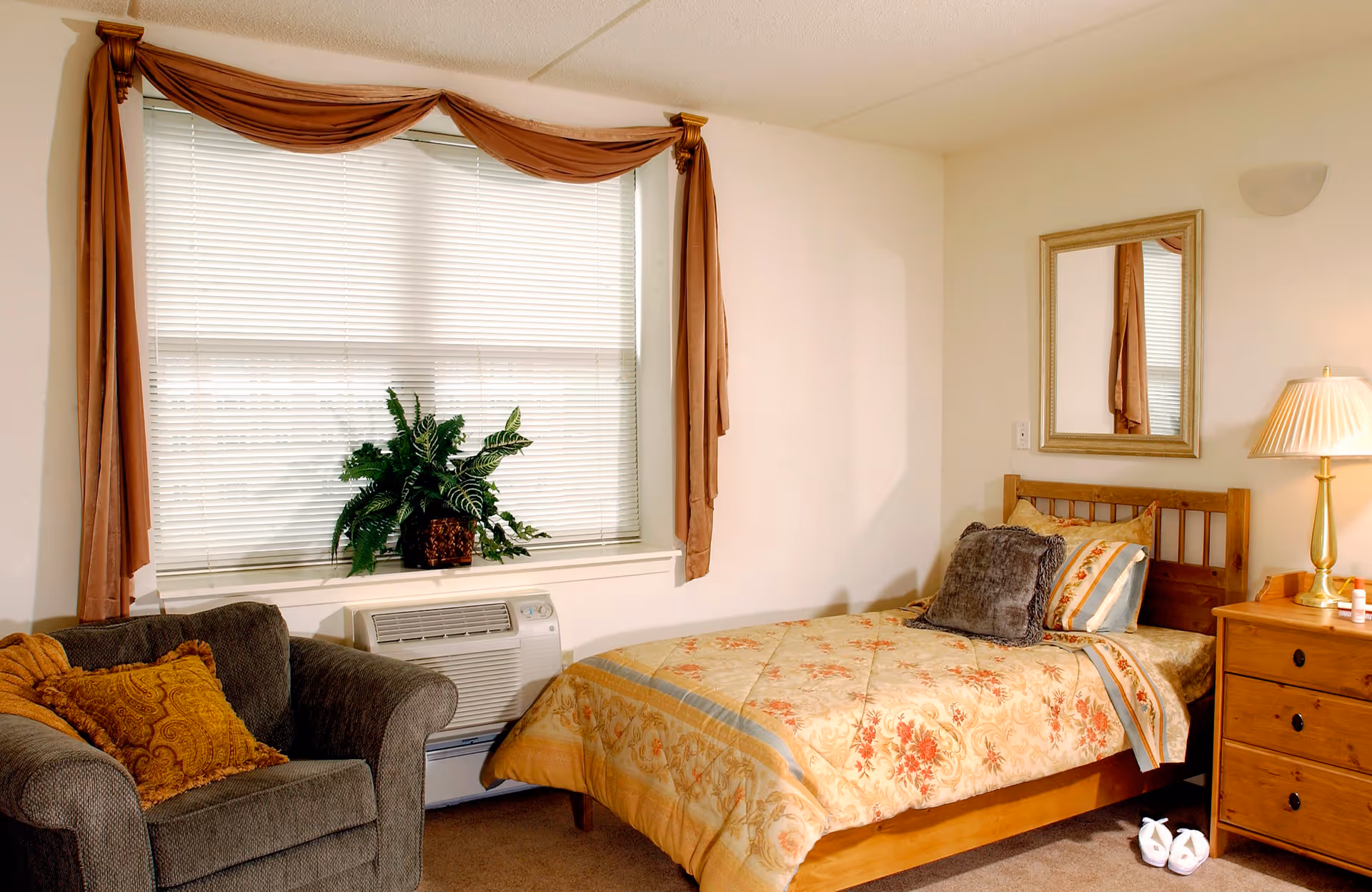 A cozy bedroom with a single wooden bed covered in a floral patterned bedspread and multiple pillows. Next to the bed is a wooden nightstand with a lamp and some small items. A comfortable armchair with a decorative pillow is placed near a window with closed blinds and brown curtains. A green potted plant sits on the windowsill above an air conditioning unit. A rectangular mirror hangs on the wall above the bed.