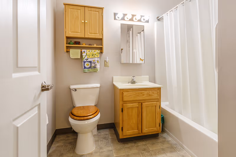 A bathroom with a white toilet featuring a wooden seat, a wooden cabinet mounted on the wall above the toilet with towels hanging beneath it, a wooden vanity with a sink and a mirror above it, and a bathtub with a white shower curtain.