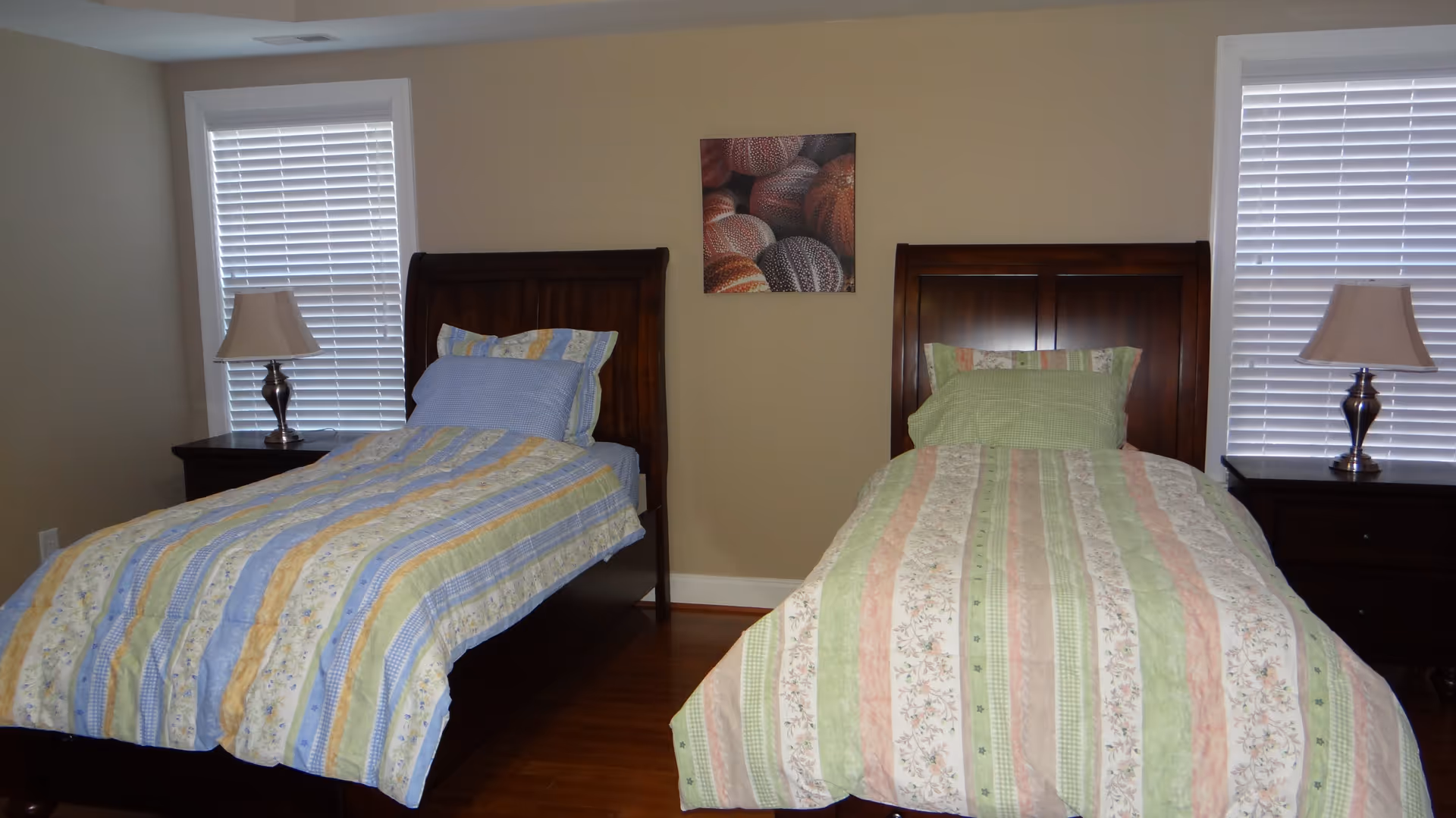 A bedroom with two twin beds, each with wooden headboards and colorful striped bedding. There are two windows with white blinds on either side of the beds, each accompanied by a dark wooden nightstand with a lamp. A picture featuring basketballs hangs on the beige wall between the beds.