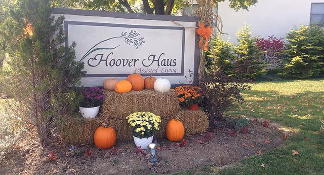 Outdoor sign for Hoover Haus Assisted Living surrounded by hay bales, pumpkins, and potted flowers in a garden area with trees and shrubs.