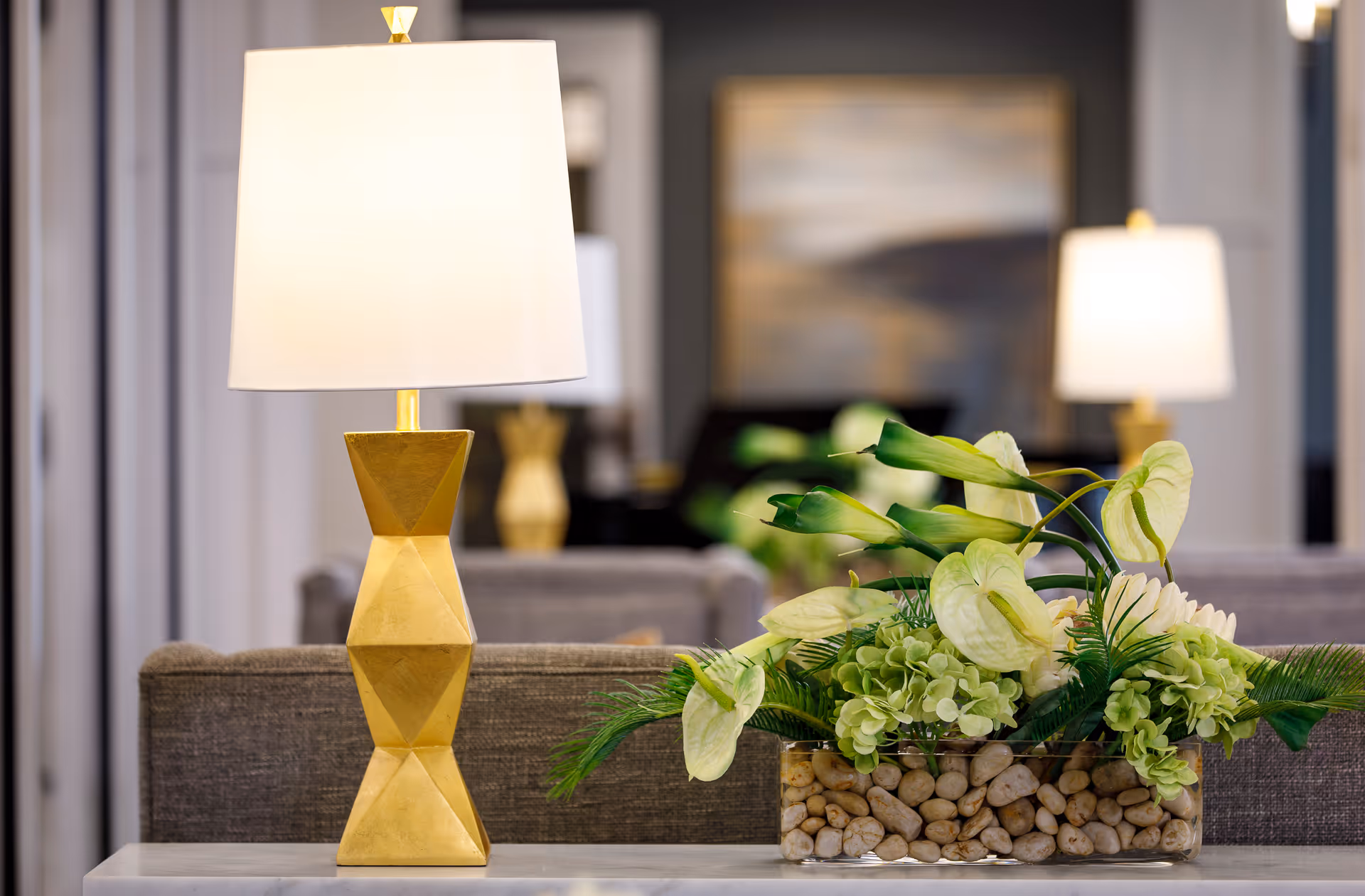 A modern table lamp with a geometric gold base and white shade sits on a table behind a sofa. Next to the lamp is a clear rectangular vase filled with decorative stones and an arrangement of green and white flowers and foliage. The background shows a softly blurred interior with another lamp and framed artwork on the wall.