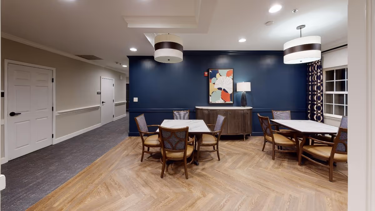 Interior view of a dining area in a senior living facility with two tables, each surrounded by four wooden chairs with cushioned seats. The room has a dark blue accent wall with a colorful abstract painting and a lamp on a wooden sideboard. Two modern pendant lights hang from the ceiling, and there is a window with patterned curtains on the right side. The floor is a light wood herringbone pattern, and a hallway with closed doors is visible on the left.