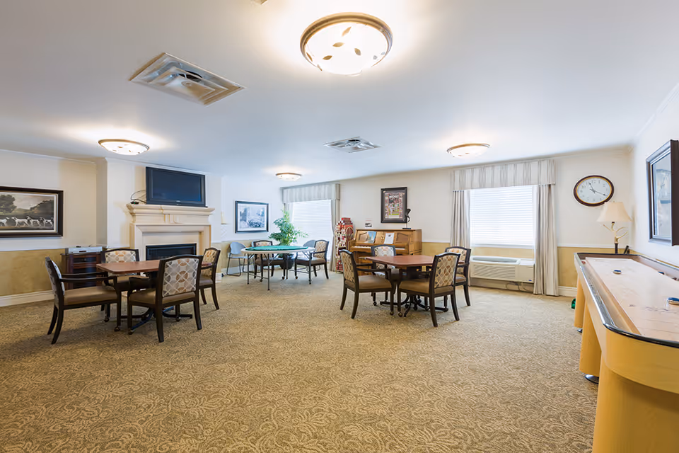 A spacious senior living common area with multiple tables and chairs arranged for socializing or activities. The room features a carpeted floor, a fireplace with a TV mounted above it, framed artwork on the walls, large windows with curtains, a clock, a lamp, and a shuffleboard table on the right side.