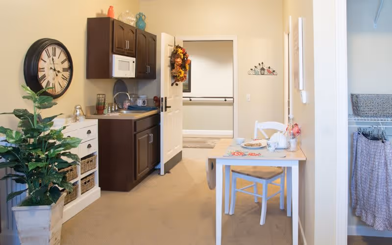 A small kitchenette area with dark wood cabinets, a microwave, and a sink. A white table with a chair is set with a teapot, cups, and plates. A large wall clock hangs on the left wall above a white storage unit with baskets. A potted plant is placed on the floor next to the storage unit. The door at the back is open, showing a hallway with handrails. On the right side, there is an open closet with hanging clothes and a basket on the shelf.