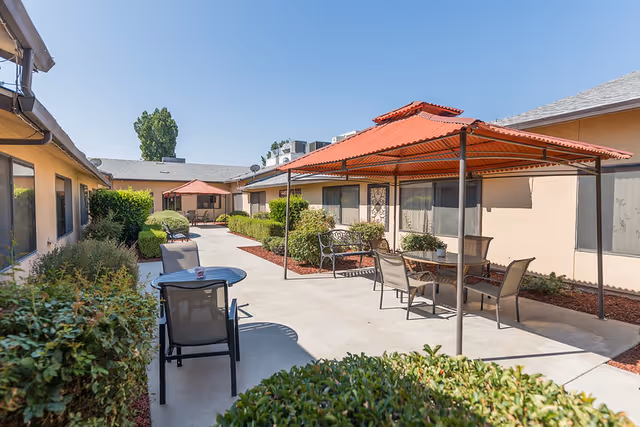 Sunlit courtyard with patio tables and chairs under red-roofed pergolas between single-story beige buildings.