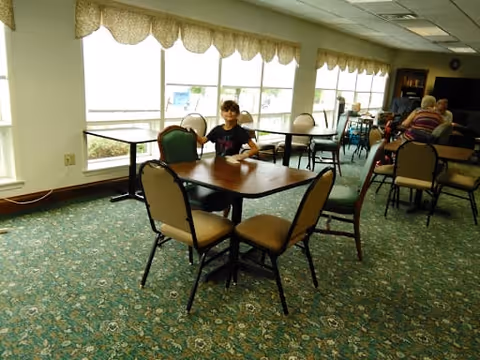 A dining area with multiple tables and chairs arranged on a patterned carpet. Large windows with valance curtains allow natural light to fill the room. A young person is seated at one of the tables, and a few elderly individuals are seated at another table in the background.