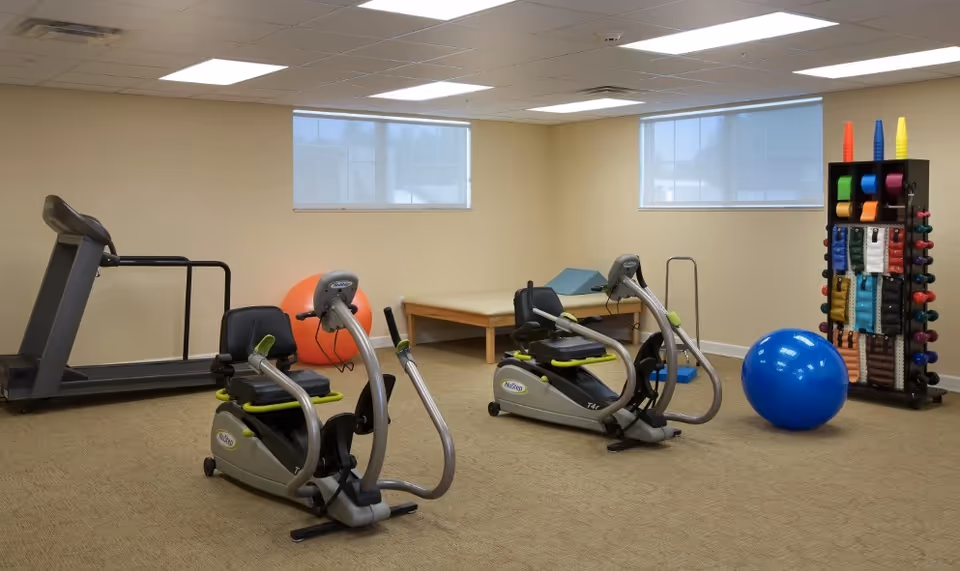 A small fitness room with exercise equipment including two recumbent exercise bikes, a treadmill, a large orange exercise ball, a large blue exercise ball, a wooden bench with a blue mat on it, and a rack holding colorful resistance bands and cones. The room has beige walls, carpeted floor, and two windows with blinds.