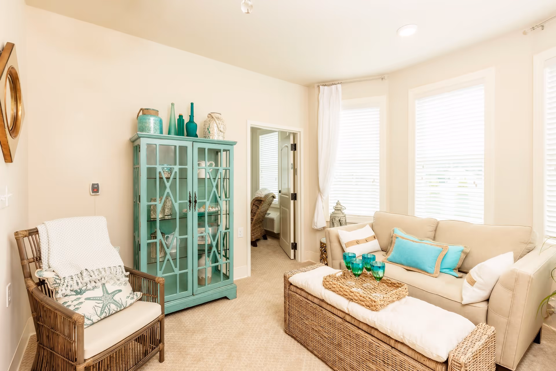 A bright living room with a beige sofa adorned with blue and white pillows, a wicker coffee table with a white cushion and a tray holding four green glasses. To the left, there is a wicker armchair with a white throw blanket and a starfish-patterned pillow. A turquoise cabinet with glass doors displays decorative items. Three large windows with white blinds and curtains allow natural light to fill the room. A doorway leads to another room with a wicker chair visible.