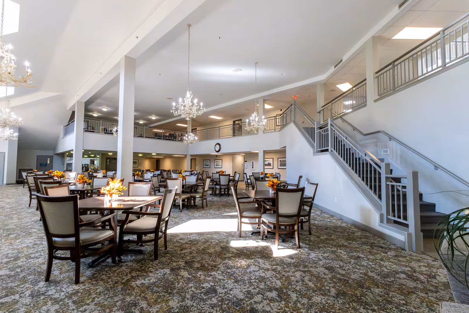 Spacious two-story dining room with round tables and chairs, chandeliers, a central staircase and a mezzanine railing.