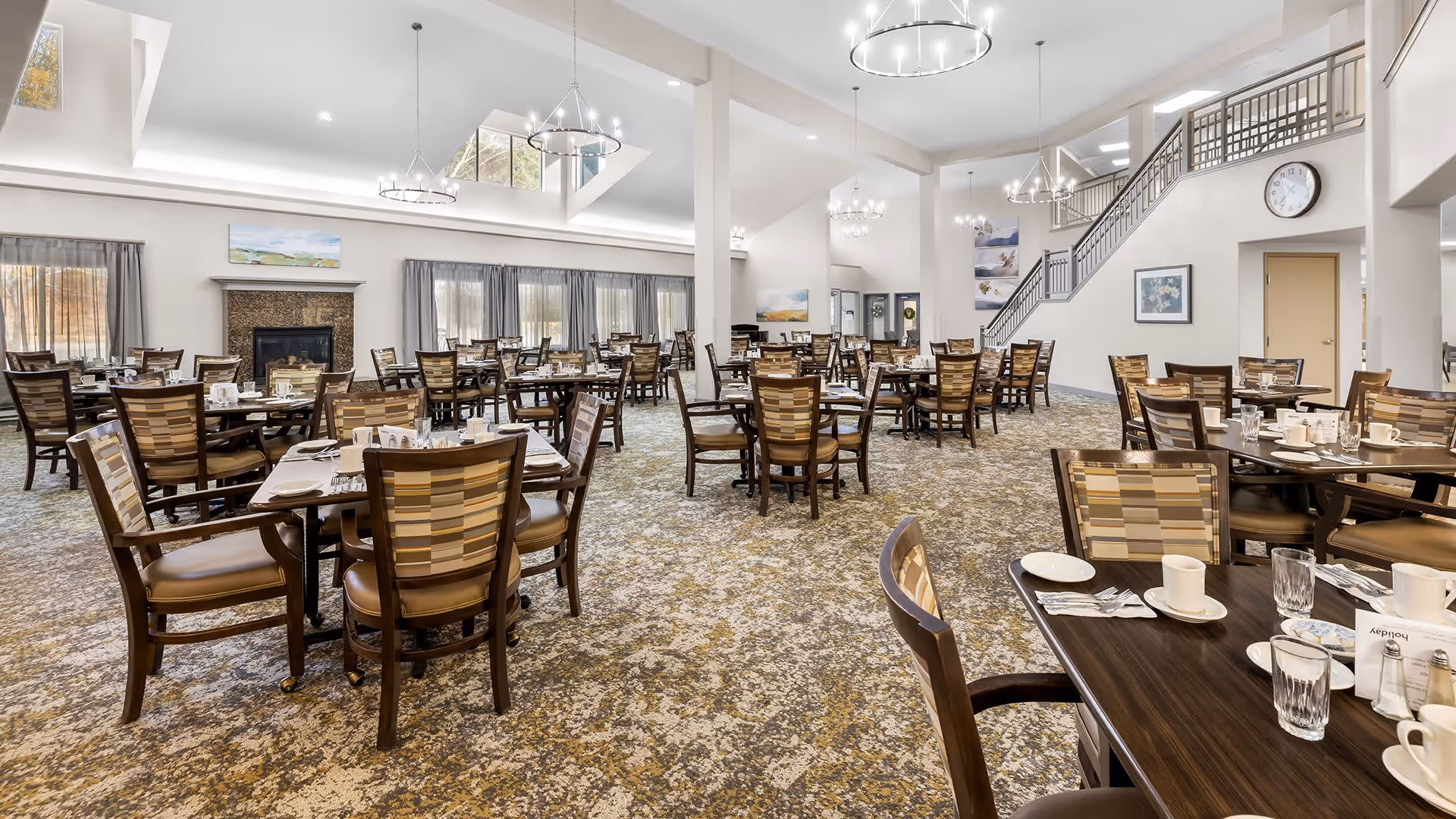 A spacious dining room with multiple wooden tables and chairs arranged neatly. Each table is set with white plates, cups, glasses, and silverware. The room has large windows with curtains, a fireplace, and modern chandeliers hanging from the ceiling. A staircase and wall clock are visible in the background.