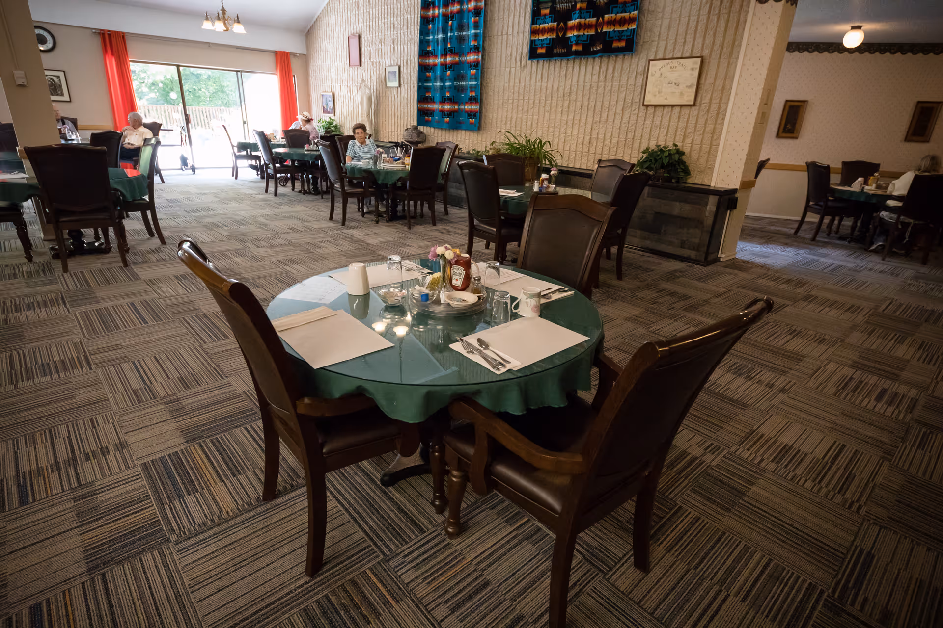 A dining room in a senior living facility with several tables covered with green tablecloths and set with placemats, utensils, glasses, and condiments. There are a few elderly residents seated at tables near large windows with red curtains, allowing natural light to fill the room. The walls have decorative textiles and framed pictures, and the floor is carpeted with a patterned design.
